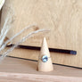 A silver ring with a round blue-gray stone displayed on a small wooden cone stand, set on a wooden shelf in front of a branded wooden box.